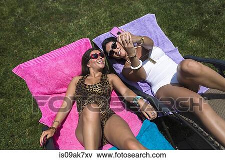 Stock Photo - Two sisters taking a selfie whilst sunbathing on loungers in garden. Fotosearch