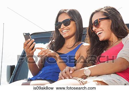 Two young women sitting on jeep hood looking at smartphone View Large Photo Image Stock Photo - Two young women sitting on jeep hood looking at smartphone. Fotosearch