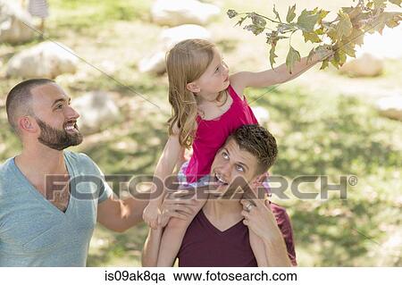 Stock Image - Young girl sitting on fathers shoulders reaching for tree leaf in park. Fotosearch