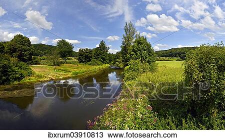 Stock Image - "Altmuhlt river flows between green fields and trees, cloudy sky, Altmuhl Valley Nature Park, Obereichstatt, Bavaria, Germany, Europe". Fotosearch