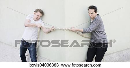 Stock Image - "Angry couple pulling a rope from opposite sides of corner of house, conceptual image of conflict in a relationship, opposing sides and determination". Fotosearch