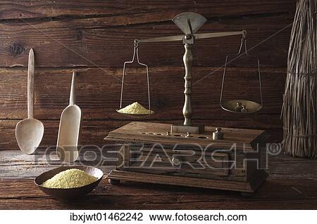 Stock Image - Antique scales weighing grains of Millet (Panicum miliaceum) on a wooden surface. Fotosearch