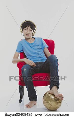 Stock Photography - "Boy, 12, sitting on a red chair wearing golden headphones with a gold football". Fotosearch