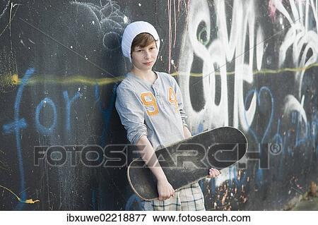 Stock Photo - Boy holding a skateboard standing in front of a wall with graffiti. Fotosearch