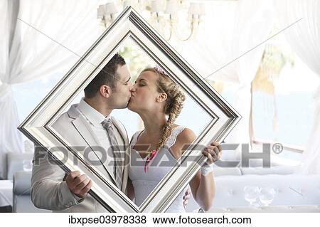 Stock Photo - "Bride and groom holding a picture frame in front of them, kissing, photo shoot". Fotosearch