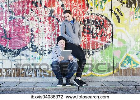 Stock Image - "Casual and relaxed young man and woman reading on a notepad in front of grafitti wall in urban area, Sweden, Europe". Fotosearch