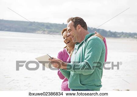 Couple playing with a digital tablet on a beach View Large Photo Image Picture - Couple playing with a digital tablet on a beach. Fotosearch