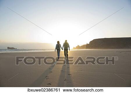 Stock Image - Couple walking along the beach towards the sunset. Fotosearch