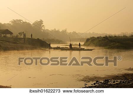 Stock Image - "Dugout canoe with punting ferryman crossing a small river early in the morning, rising sun and fog, Chitwan National Park, Nepal, Asia". Fotosearch