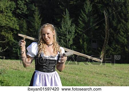 Stock Image - "Farm girl with a scythe, wearing a dirndl, a traditional German or Austrian dress". Fotosearch