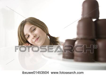 Picture - Girl sitting mischievously in front of a plate of chocolate marshmallows. Fotosearch