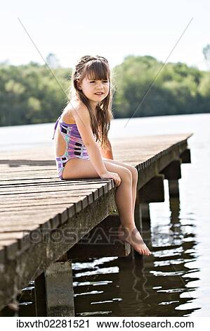Stock Image - Girl sitting on jetty at lakeside. Fotosearch