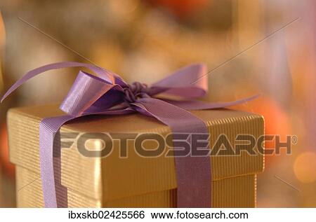 Stock Photograph - "Golden gift box with a purple ribbon, in front of a Christmas tree". Fotosearch
