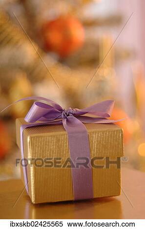 Stock Photography - "Golden gift box with a purple ribbon, in front of a Christmas tree". Fotosearch