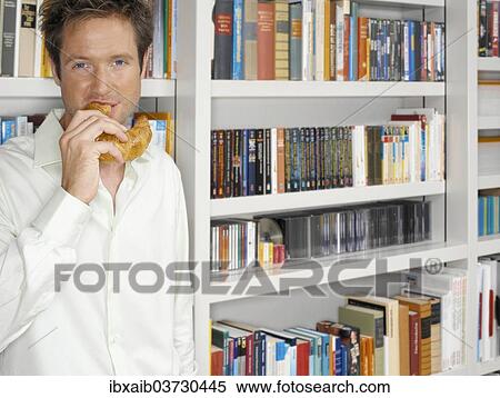 Stock Photography - Man standing in front of a bookshelf and eating a croissant. Fotosearch