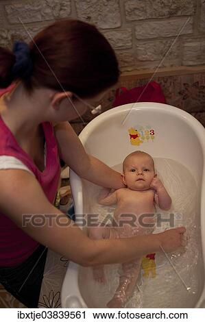 Stock Image - "Mother bathing her baby girl, 2 months, in a baby bathtub". Fotosearch