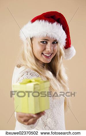 Stock Image - Smiling blond young woman wearing a Santa hat and presenting a gift. Fotosearch