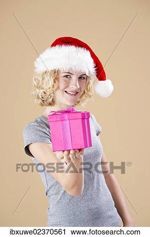 Stock Image - Smiling blond young woman wearing a Santa hat and presenting a gift. Fotosearch