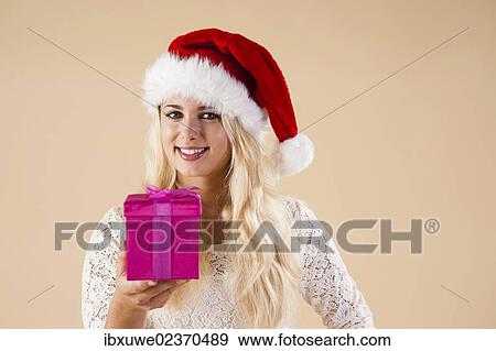Smiling blond young woman wearing a Santa hat and presenting a gift View Large Photo Image Stock Photo - Smiling blond young woman wearing a Santa hat and presenting a gift. Fotosearch