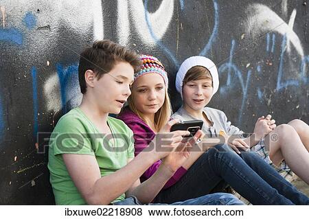 Three teenagers with a Smartphone sitting in front of a wall with graffiti View Large Photo Image Stock Photo - Three teenagers with a Smartphone sitting in front of a wall with graffiti. Fotosearch