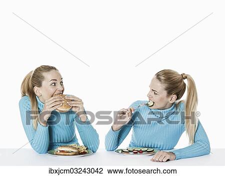 Stock Image - "Two women eating food, one eating fast food, the other one eating uncooked vegetarian food, Germany, Europe". Fotosearch