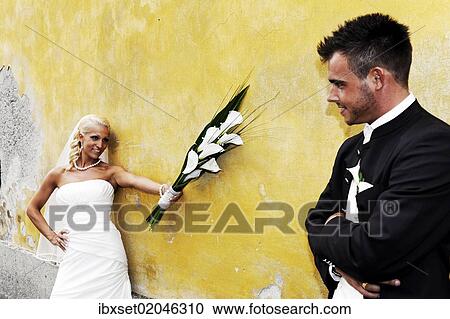 Stock Image - "Wedding, laughing bride holding out the bridal bouquet towards the groom in front of a yellow wall". Fotosearch