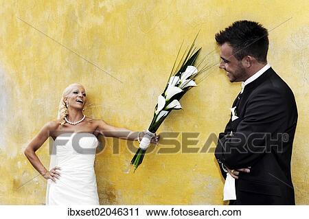 Stock Image - "Wedding, laughing bride holding out the bridal bouquet towards the groom in front of a yellow wall". Fotosearch