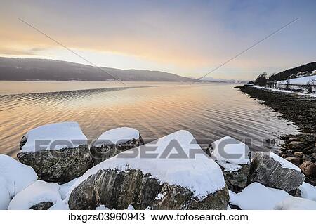 Stock Image - "Winter atmosphere at sunrise on the fjord, snow-covered rocks, coastal road, near Alesund, Norway, Europe". Fotosearch