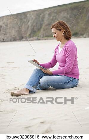 Stock Photograph - Woman sitting with a digital tablet on a beach. Fotosearch