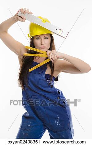 Stock Image - Woman wearing blue overalls and a hardhat holding a folding carpenter's ruler and a spirit level. Fotosearch