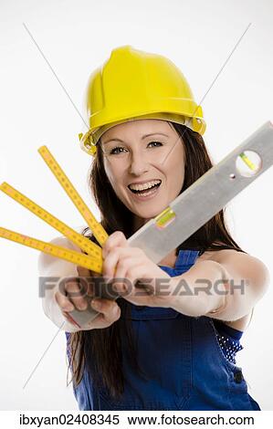 Stock Photography - Woman wearing blue overalls and a hardhat holding a folding carpenter's ruler and a spirit level. Fotosearch