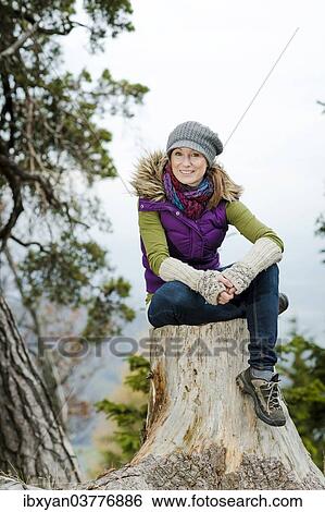 Stock Photograph - Woman wearing winter clothes sitting on a tree stump. Fotosearch