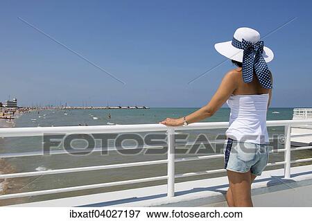 Stock Photo - "Woman with hat standing at railing on the beach, Adriatic, Senigallia, Province of Ancona, Marche, Italy, Europe". Fotosearch