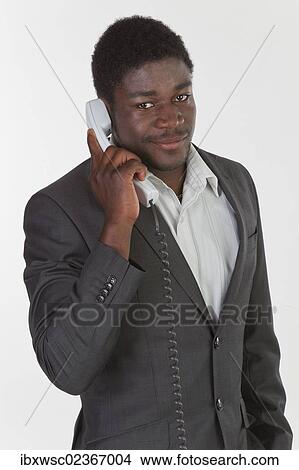 Picture - "Young black man in a suit making a phone call in an office, smiling". Fotosearch
