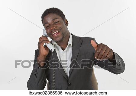 Stock Photograph - "Young black man in a suit making a phone call in an office, smiling with a thumbs up gesture". Fotosearch