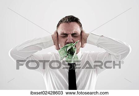 Stock Image - "Young, desperate man wearing a shirt and a tie holding his hands over his ears, has 100-euro notes stuffed into his mouth". Fotosearch