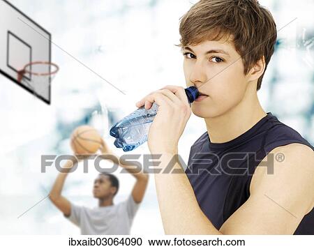 Stock Image - "Young man, holding a water bottle, in front of a young man with a basketball in a sports hall". Fotosearch