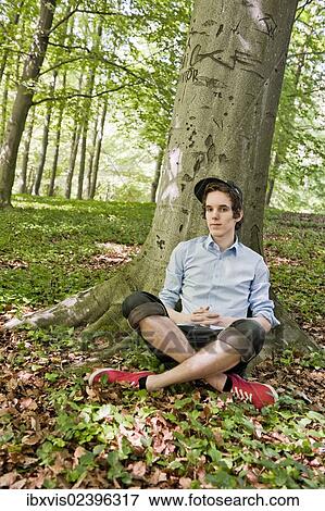Stock Photo - "Young man sitting in front of a tree in the woods, Sweden, Europe". Fotosearch