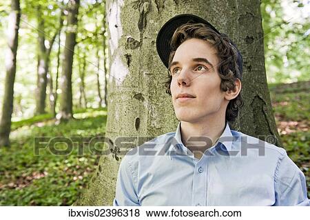Stock Photo - "Young man sitting in front of a tree in the woods, Sweden, Europe". Fotosearch