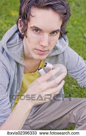 Stock Image - "Young man sitting in nature, holding lightbulbs". Fotosearch