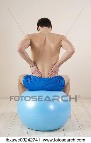 "Young man sitting on a gym ball and touching his aching back, Mannheim, Baden-Wurttemberg, Germany, Europe" View Large Photo Image Stock Image - "Young man sitting on a gym ball and touching his aching back, Mannheim, Baden-Wurttemberg, Germany, Europe". Fotosearch