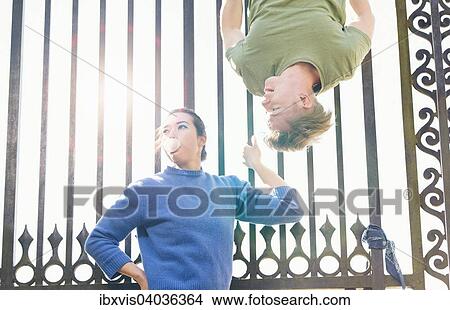 Picture - "Young man trying to impress indifferent girl by hanging upside down on an iron gate, Sweden, Europe". Fotosearch