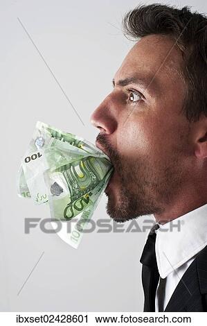 Stock Image - Young man wearing a suit with 100-euro notes stuffed into his mouth. Fotosearch