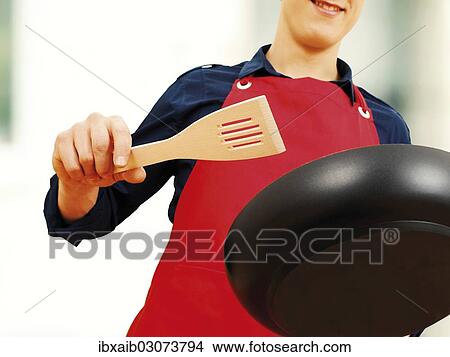 Young man wearing an apron holding a pan and a spatula View Large Photo Image Picture - Young man wearing an apron holding a pan and a spatula. Fotosearch