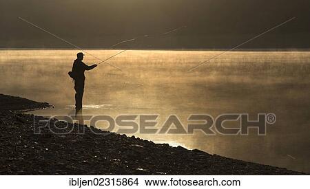 Picture - "A fly fisherman standing on the shore of the Obernau reservoir at sunrise, Netphen, North Rhine-Westphalia, Germany, Europe". Fotosearch