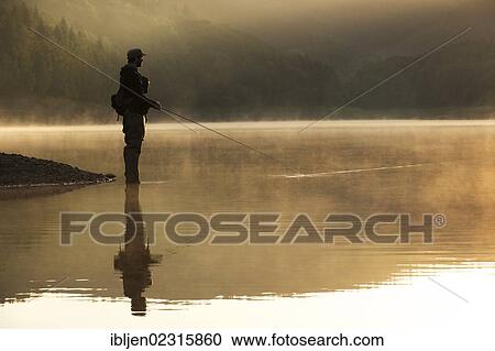 Stock Image - "A fly fisherman standing on the shore of the Obernau reservoir at sunrise, Netphen, North Rhine-Westphalia, Germany, Europe". Fotosearch