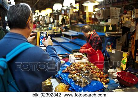 Stock Image - "A man uses a smart phone to take a photograph at the Noryangjin Fish Market, Seoul, South Korea, Asia". Fotosearch