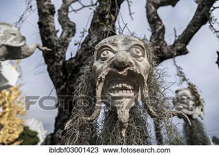 Stock Image - "A mask hanging on a tree, at the Wat Rong Khun temple or The White Wat, a Buddhist-Hindu temple, Chiang Rai, Chiang Rai Province, Northern Thailand, Thailand, Asia". Fotosearch