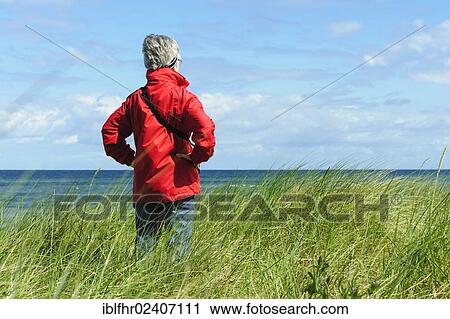 Stock Image - "A woman standing in the dunes and looking out on the sea, Poel island, Mecklenburg-Western Pomerania, Germany, Europe". Fotosearch