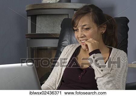 Stock Image - "A young woman of Indonesian origin is sitting in front of a wood burner working on a laptop computer, Germany, Europe". Fotosearch